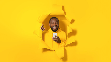 Happy black man holding modern cellular and smiling at camera, posing in ripped torn yellow paper background
