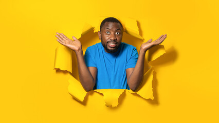 African american man spreading hands while complaining, posing in torn yellow paper hole