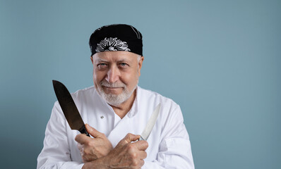 Elderly charismatic male chef in uniform holding two knives, looking at camera and smiling on light blue background, vertical format, space for text