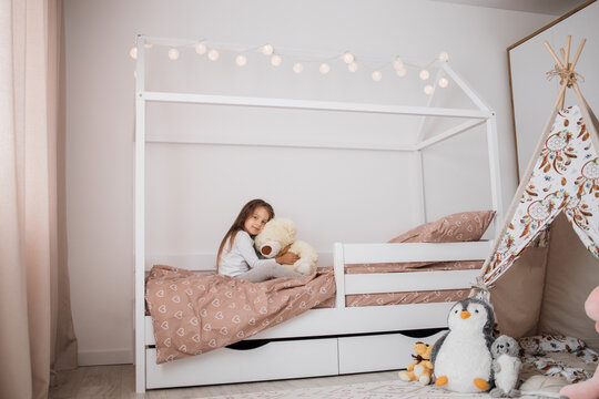 Cute little girl in pajama hugging her toy bear on the scandinavian wooden house frame bed at home, happy childhood concept, indoor horizontal portrait.
