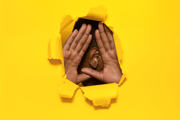 African american man holding hands near ear, listening through torn paper yellow background