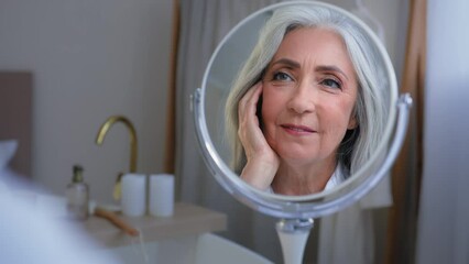 Pensive 50s middle-aged Caucasian woman senior touching face skin looking at mirror reflection smiling enjoy soft facial. Older lady pampering healthy skincare beauty cosmetology rejuvenation