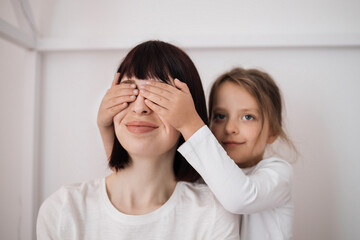 Satisfied caucasian preschooler girl closes eyes to young lady and congratulates with birthday, sitting on bed in bedroom interior. Surprise, holiday, love and relationship. Celebrate at home