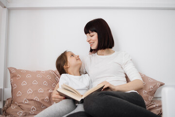 Close up of smiling mom and little daughter relax in cozy bed read funny interesting children book...