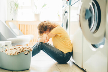 Depressed woman sitting near washing machine at laundry.