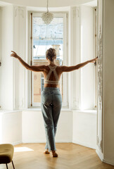 A beautiful slim woman in jeans and a bra stands with her back to the camera and looks out the window