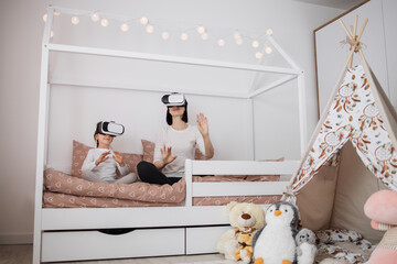 New digital technology. Joyful nice caucasian mother and daughter smiling while using virtual reality glasses at home sitting on scandinavian wooden bed.