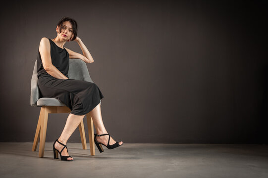 Studio portrait of young beautiful sensual woman in blak suit sitting on wooden chair against dark background.