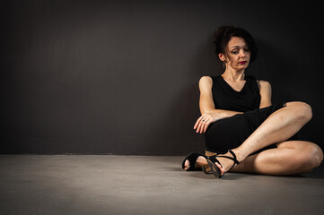 Fototapeta na wymiar Studio portrait of a tired, tortured, young beautiful sensual woman in a black dress sitting on the floor on a gray background. Depression, fatigue, loneliness, thoughtfulness