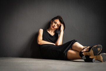 Studio portrait of a tired, tortured, young beautiful sensual woman in a black dress sitting on the floor on a gray background. Depression, fatigue, loneliness, thoughtfulness