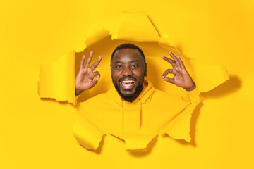 Everything is under control. Happy black man giving positive opinion, making ok gesture posing in torn yellow paper hole - obrazy, fototapety, plakaty