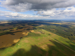 Aerial panorama drone view of typical agricultural landscape. Green farmers field and forest. Cloudy skies. A sunny autumn day. Photo above shadows from the clouds on the ground.