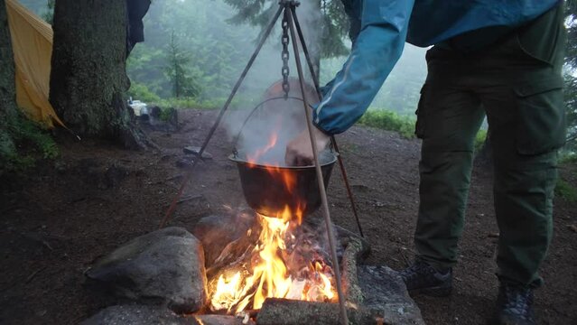Cooking on a fire in the forest during the rain. Cooking on a camping trip in the mountains. Lifting the lid of the cauldron and stirring the food with a spoon. Hiking and tourist lunch. Footman. 4k