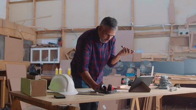 Mid adult man talking to client on mobile phone while working in woodworking factory