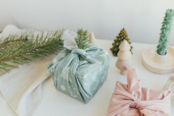 Furoshiki gift wrapping. Stylish wrapped gifts in linen fabric on white rustic table with eco friendly wooden toys, fir branches, candle. Zero waste Christmas concept. Happy Holidays!