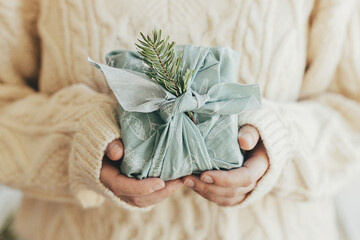 Hands holding gift wrapped in green fabric with fir branch. Stylish Furoshiki gift. Woman in...