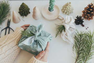 Zero waste Christmas. Hands holding gift box wrapped in green fabric with fir branch on white wooden table with eco friendly decor and candle. Furoshiki gift wrapping. Merry Christmas!