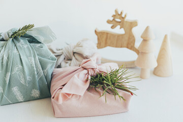 Stylish wrapped gifts in linen fabric on white rustic table with eco wooden tree, deer, fir branches, candle. Furoshiki gift wrapping. Eco friendly toys.  Zero waste Christmas. Merry Christmas!