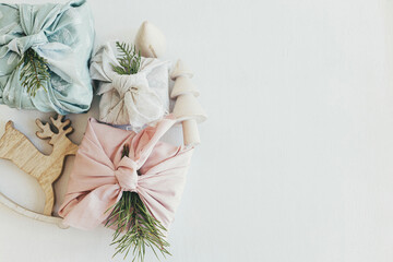 Christmas flat lay. Stylish wrapped gifts in linen fabric on white rustic table with eco wooden tree, deer, fir branches, candle. Space for text. Furoshiki gift wrapping. Zero waste