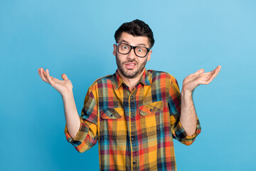Photo of handsome young confused guy in eyewear dressed plaid shirt question shrugging shoulders isolated on blue color background
