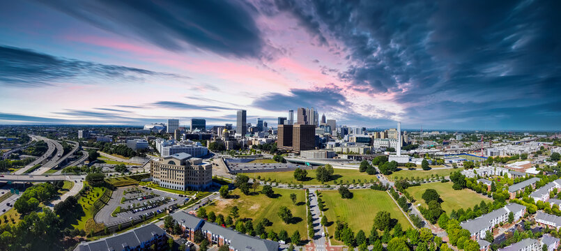 An Aerial Panoramic Shot Of The Skyscrapers And Office Buildings In The City Skyline With Vast Miles Of Lush Green Trees, Grass And Plants And Cars On The Street With Powerful Clouds At Sunset