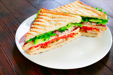 Homemade club sandwich with toasted bread slices, chicken breasts, tomato, salad leafs, cucumber, onion on white plate. On wooden tabletop. Side view.