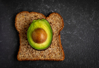 A half of an avocado with a seed sitting on a piece of whole wheat toast on a granite countertop