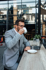 Middle age businessman sitting in cafe bar and enjoying at coffee break between two meetings. He uses laptop computer and he is positive and confident.