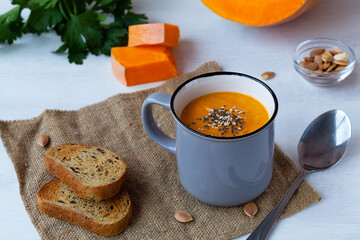 Pumpkin cream soup in a grey cup with pieces of pumpkin with green on a white kitchen table. Mug of tasty Pumpkin soup with seeds and crackers on the sackcloth. Vegetarian autumn soup. copy space.