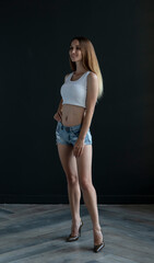 young woman in a white tank top and denim shorts on a dark background, full length portrait