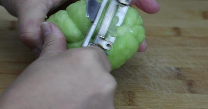 Chayote on the wooden chopping board and adult female hands starting to peel with a peeler, known as: labu siam, jipang, organically grown vegetables. Healthy homemade food concept