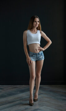 young woman in a white tank top and denim shorts on a dark background, full length portrait