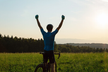 Male racing cycle rider celebrating the win with arms raised above his head, on a hill surrounded...