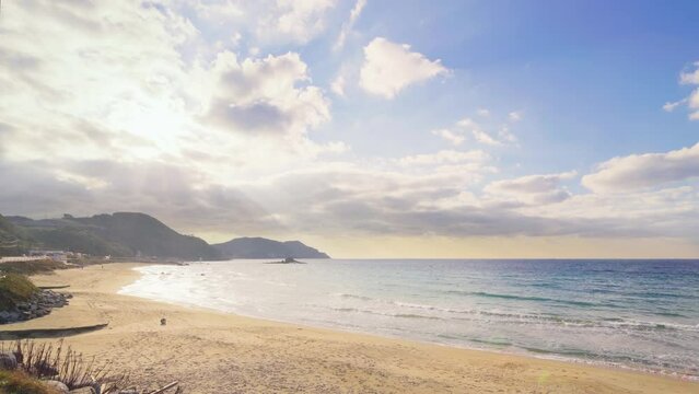 Wide angle video of the coast of the empty Itoshima beach with in background the Sakurai Futamigaura Meoto Iwa Couple Rock and its white torii gate lighted by sunset light filtering through clouds.