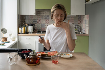 Mature woman tastes yummy jam making toast with butter. Middle aged female enjoys preparing tasty...
