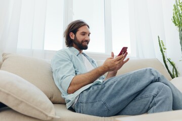 A man with a beard sits on the couch during the day and smiles at home and looks at his phone relaxing on his day off, life online on his phone texting with friends