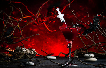 Halloween alcoholic cocktail bloody martini with syringe on scary dark red background with twisted branches, bats, stones, pumpkin guards and spiders, festive drink for vampire party
