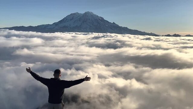 man stands on a mountain above the clouds with his arms outstretched before the world.