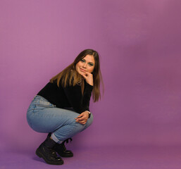 full length shot of a beautiful woman in makeup sitting on squat's position touching her chin and knee ahead a purple background