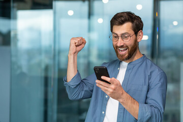 Successful businessman working in modern office building, man looking at smartphone and reading news celebrating happy news victory businessman in shirt and glasses holding hand up triumph gesture.