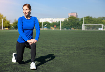 Women and sport. Girl in sportswear does exercises: kneels and stretches on the grass at the stadium on a sunny day. Middle aged sportswoman dressed in sportsclothes exercising outdoors