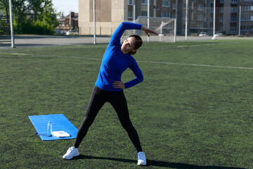 Women and sport. Girl in sportswear does exercises: bends and stretches on the grass at the stadium on a sunny day. Middle aged sportswoman dressed in sportsclothes exercising outdoors