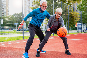 Cheerful active senior couple playing basketball on the urban basketball street court. Happy living...