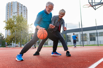 Cheerful active senior couple playing basketball on the urban basketball street court. Happy living after 60.