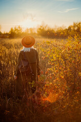 Fototapeta premium Rear, back view of fashionable woman wearing orange hat, olive green trench coat, with leather backpack, posing in beautiful autumn nature during the sunset. Copy, empty space for text