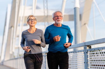 Cheerful active senior couple jogging together outdoors on the bridge. Healthy activities for...