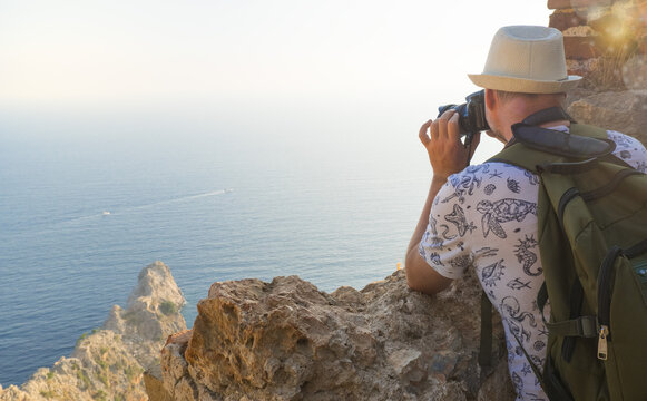 A young man with a backpack takes pictures of the landscape on top of a mountain. Back view.