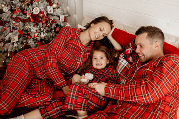 Dad, mom and daughter in red pajamas lie on the bed and enjoy the Christmas holidays. Beautifully decorated house in Christmas decor. The family rejoices and has fun lying on the bed