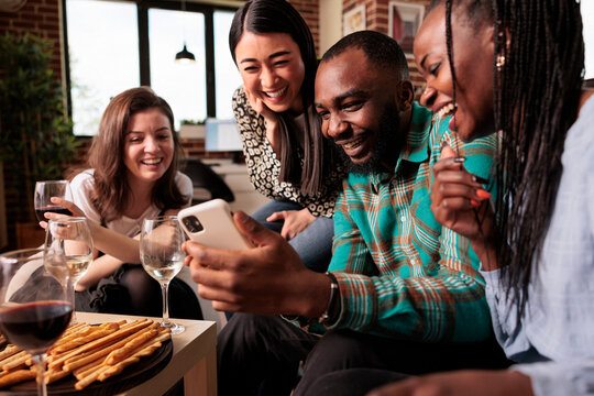 Happy multiple nationalities, ethnicities group friends looking, watching funny messages at smartphone screen, smiling, laughing, drinking wine, eating bread sticks at apartment reunion.