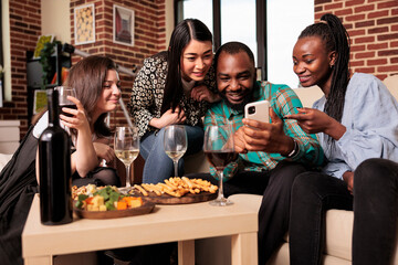 Diverse nationality group people staring at smartphone screen, smiling, laughing, enjoying friendship, drinking alcohol, wine, tasting cheese, bread sticks at home made party.